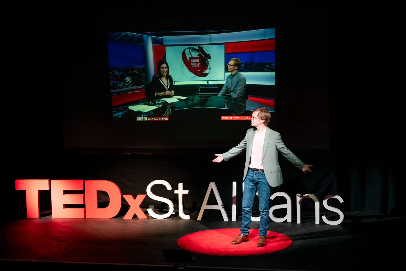 Nat Hawley speaking at TEDx St Albans, with a BBC World News clip on the screen behind