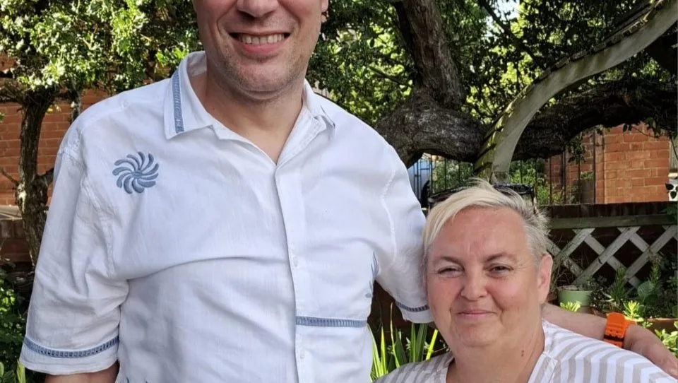 Adem Ahmet, in a white shirt, standing beside a woman with short blonde hair under a tree