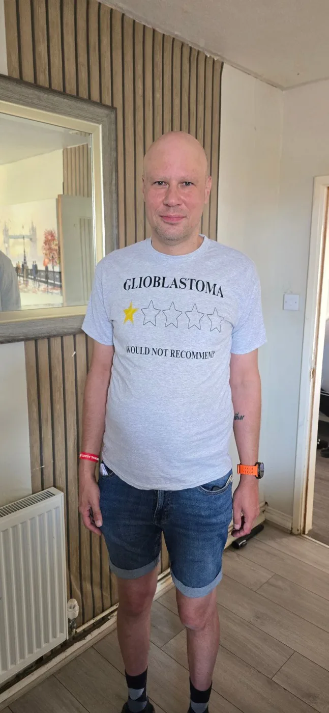 Adem Ahmet standing in a doorway in a white T-shirt printed with the word Glioblastoma, one star out of five and the words Would not recommend