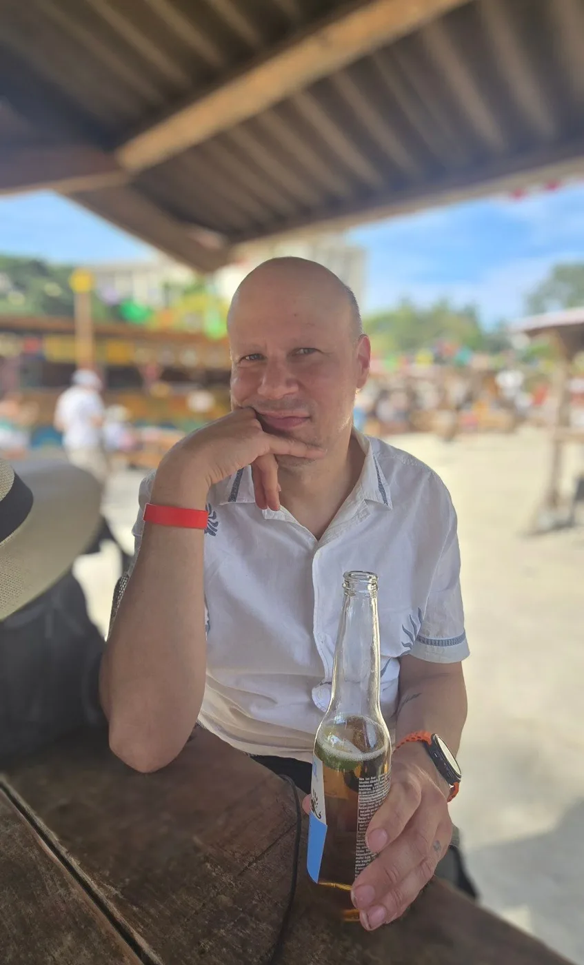 Adem Ahmet sitting at an outdoor bar table in a white shirt, holding a bottle, with a thatched shade behind him