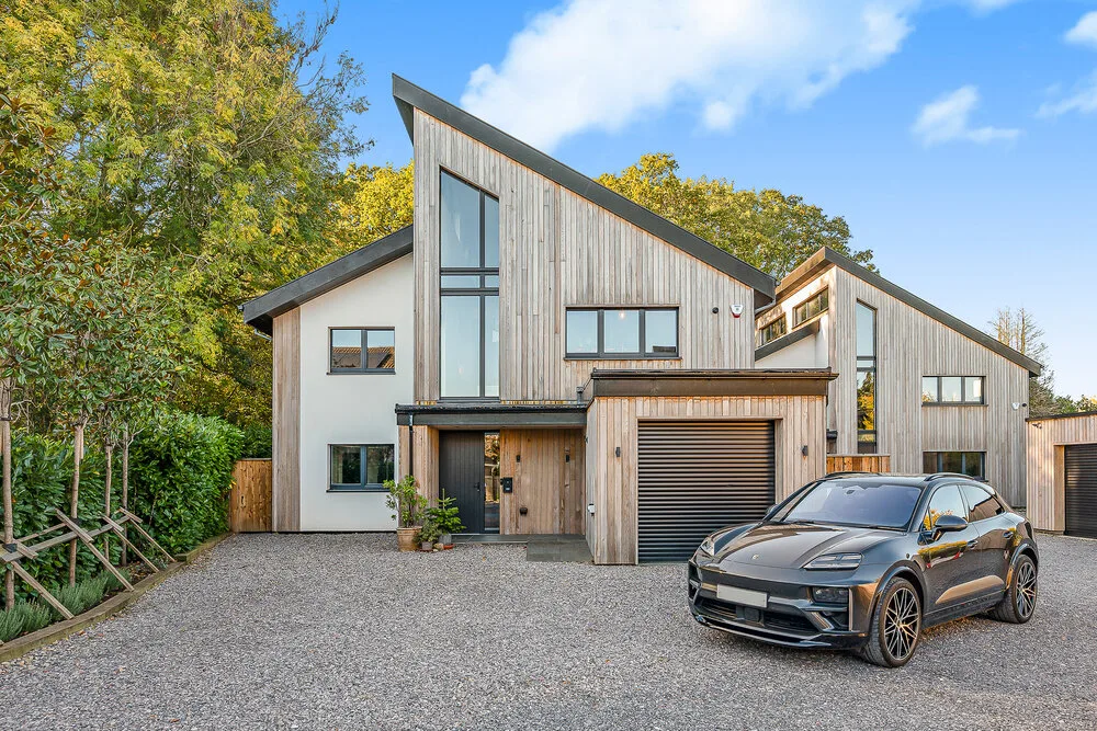 Two timber-clad modern houses with steep asymmetric roofs, tall windows and a garage door, on a gravel drive with trees behind