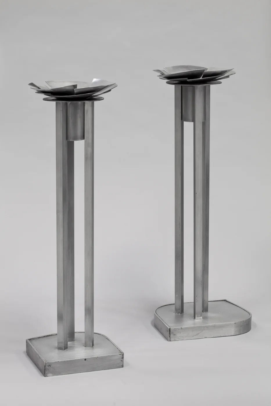 Two tall metal candlesticks, each with square legs and a flat petal-like top, photographed against a pale grey background