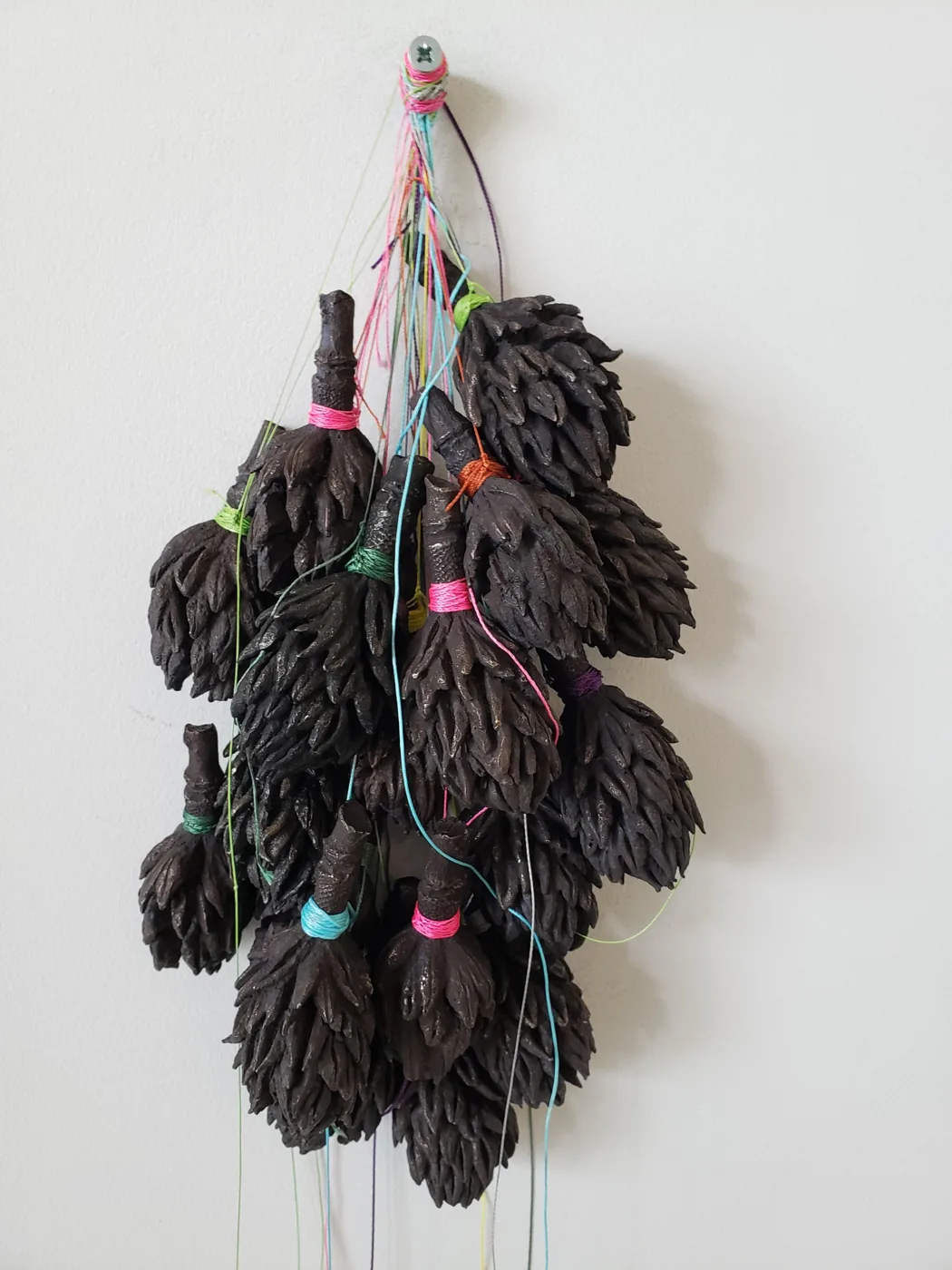A hanging cluster of dark, leaf-like sculpted forms bound with pink, green, orange and blue threads, against a white wall