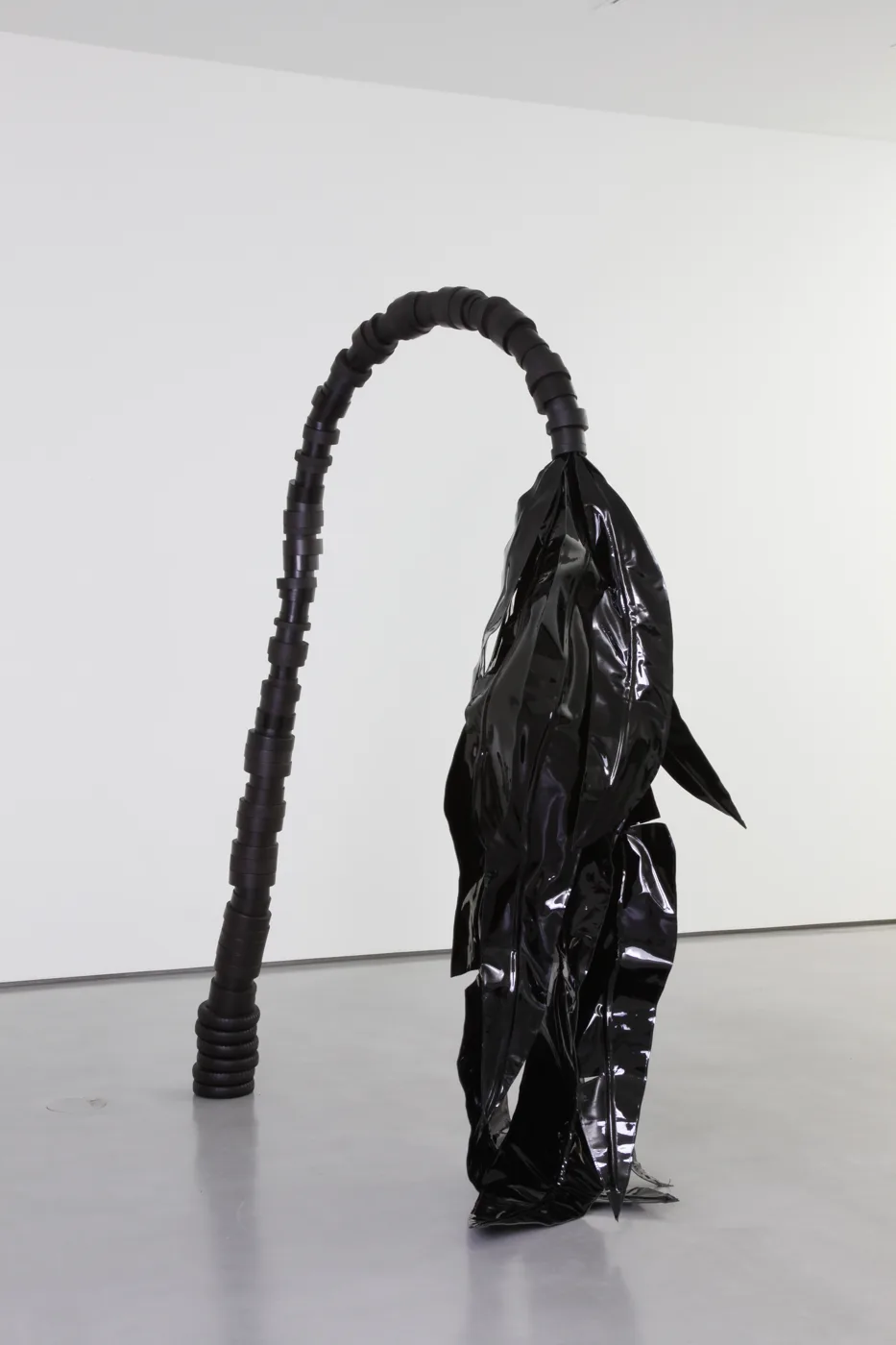 A tall black sculpture in a white gallery: a ridged black arch curving over into a draped shape of glossy black material, standing on a grey floor